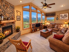 A cozy living room with a fireplace, large windows with mountain views, a ceiling fan, and a comfortable seating area.