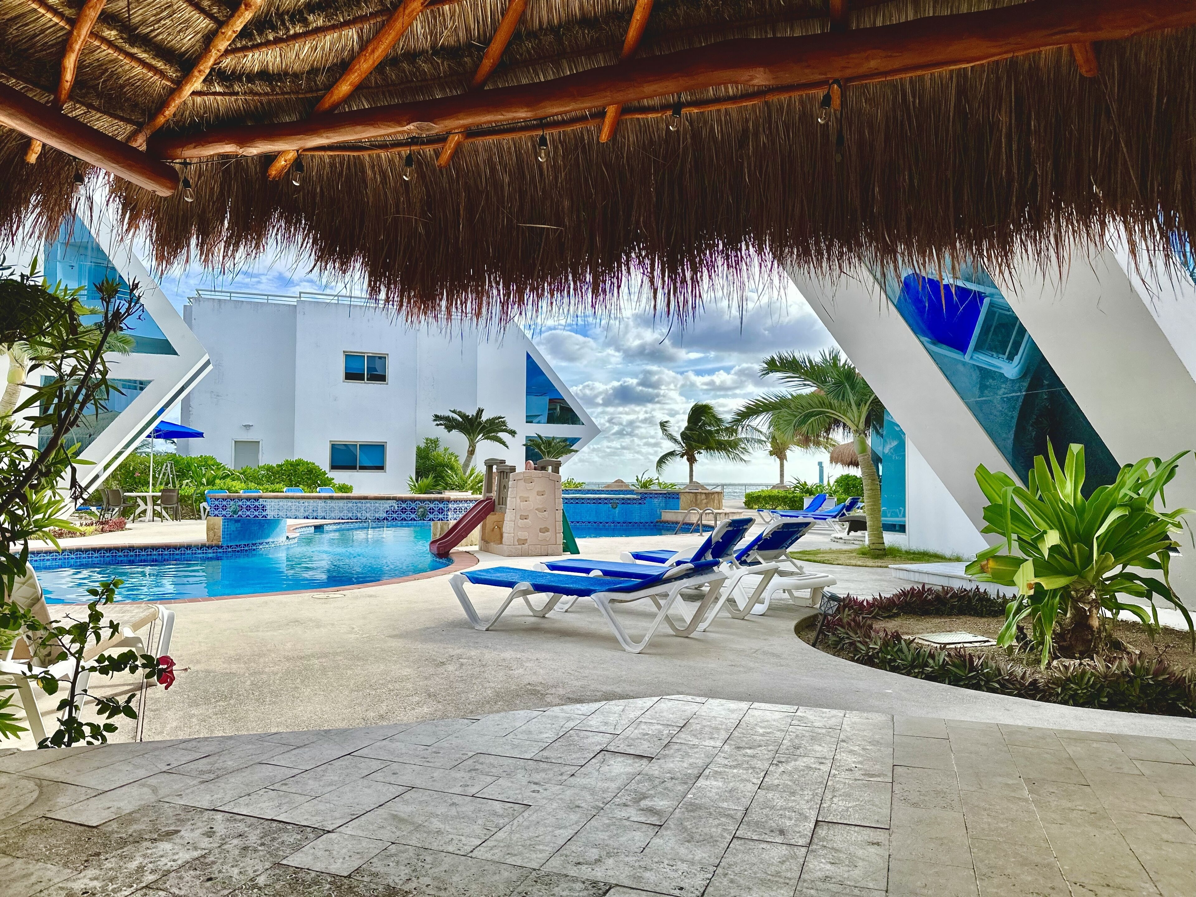 A pool area with lounge chairs, a slide, and a thatched roof structure.