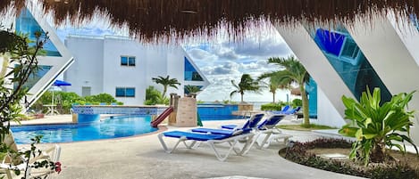 A pool area with lounge chairs, a slide, and a thatched roof structure.