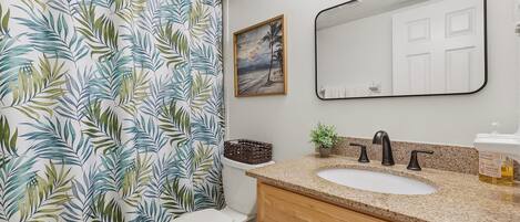 Un baño con inodoro, lavamanos, espejo y cortina de ducha estampada con hojas.