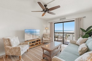 A living room with a television, a ceiling fan, a sofa, a chair, and a view of the beach through a sliding door. - 410 - Boho Fourth Floor 1/1 Sleeps 2 (New Smyrna Beach)