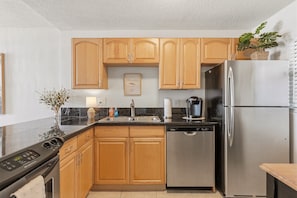 A kitchen with wooden cabinets, a stainless steel refrigerator, a dishwasher, and a coffee maker. - 410 - Boho Fourth Floor 1/1 Sleeps 2 (New Smyrna Beach)