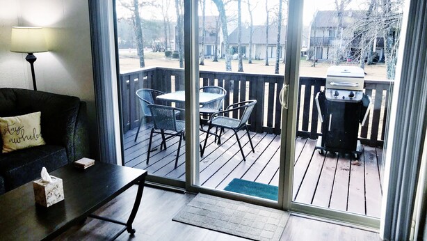 A room with a sliding glass door leading to an outdoor deck. Inside, there is a dark-colored sofa with a cushion, a small table with a tissue box, and a lamp. The deck has a grill and outdoor chairs. - 🌟Updated Fairfield Glade Condo 🌟w/King Size bed, pet friendly, Wifi, & more! (Crossville)