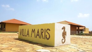 Placa para "Villa Maris" com desenho de cavalo-marinho, com um cenário de edifícios com telhados de barro vermelho.