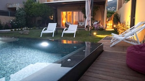 A modern outdoor pool area with a wooden deck, two white lounge chairs, and a glass-enclosed hot tub.