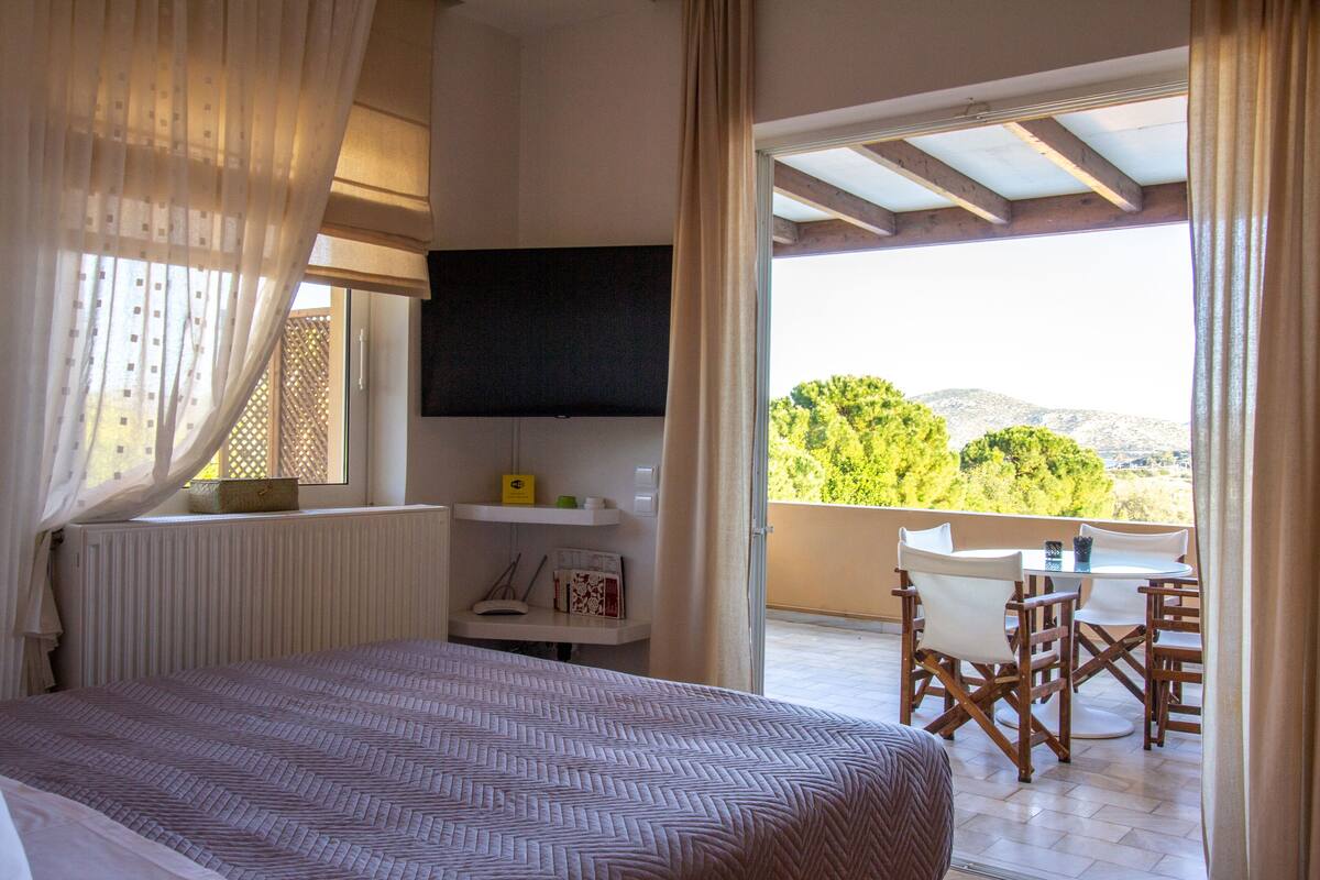 A bedroom with a bed, a TV mounted on the wall, a small table with chairs, and a balcony with a view of trees and hills.