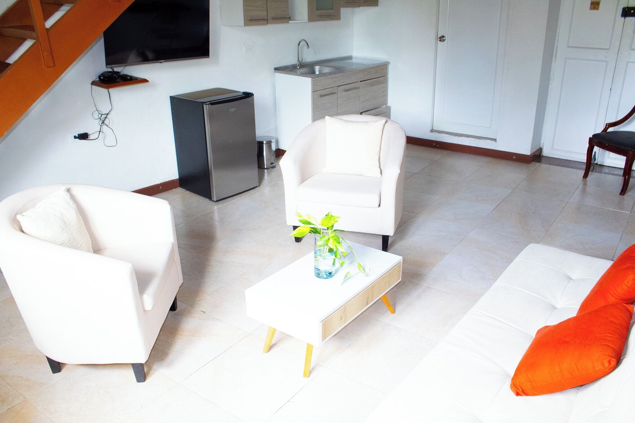 Ein modernes Wohnzimmer mit einem weißen Sofa, einem weißen Sessel und einem kleinen weißen Couchtisch mit einer Pflanze. Im Hintergrund ist eine Küchenzeile mit Spüle und Kühlschrank zu sehen.