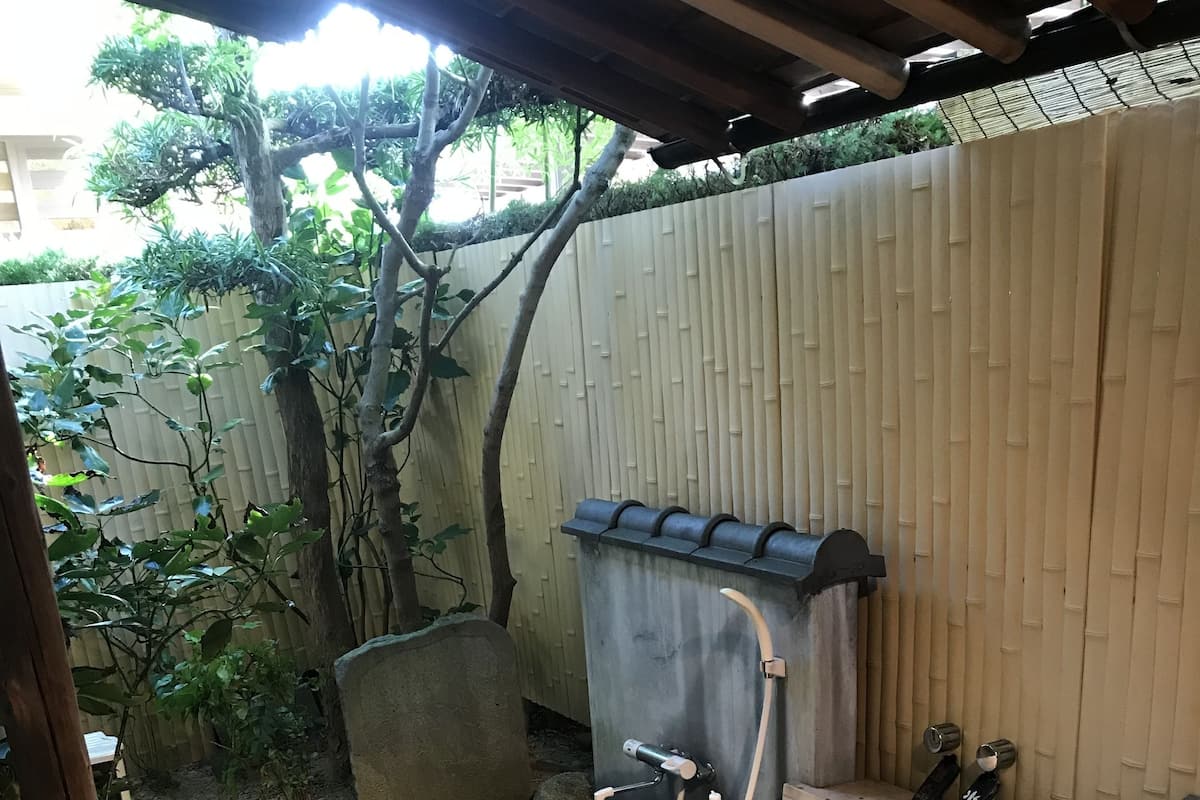 Una bañera de madera con un banco de madera, una pared de piedra y una valla de bambú al fondo.