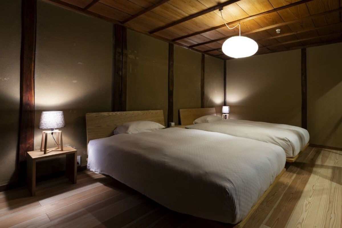 A room with two beds, wooden flooring, a ceiling light, and a nightstand with a lamp.