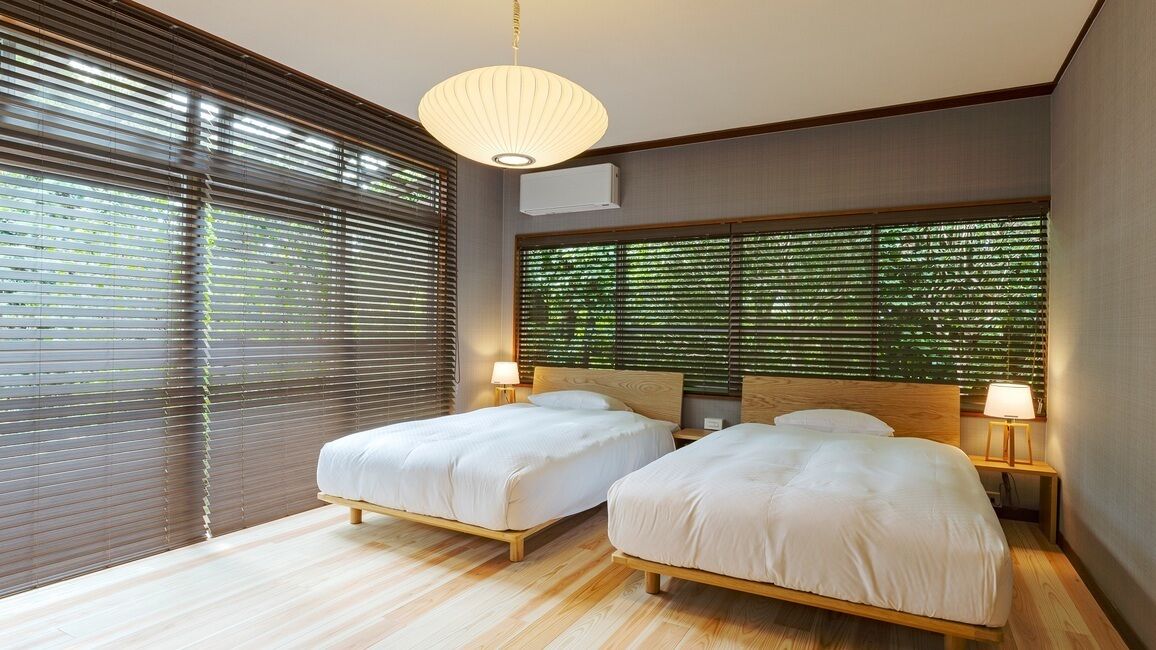 Un dormitorio moderno con dos camas, cabeceros de madera, un ventanal con persianas y una lámpara colgante.