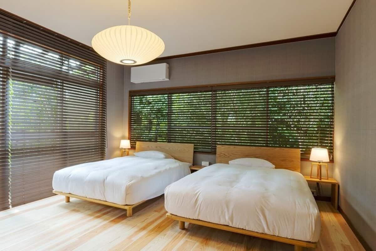 A modern bedroom with two beds, wooden headboards, a large window with blinds, and a hanging light fixture.