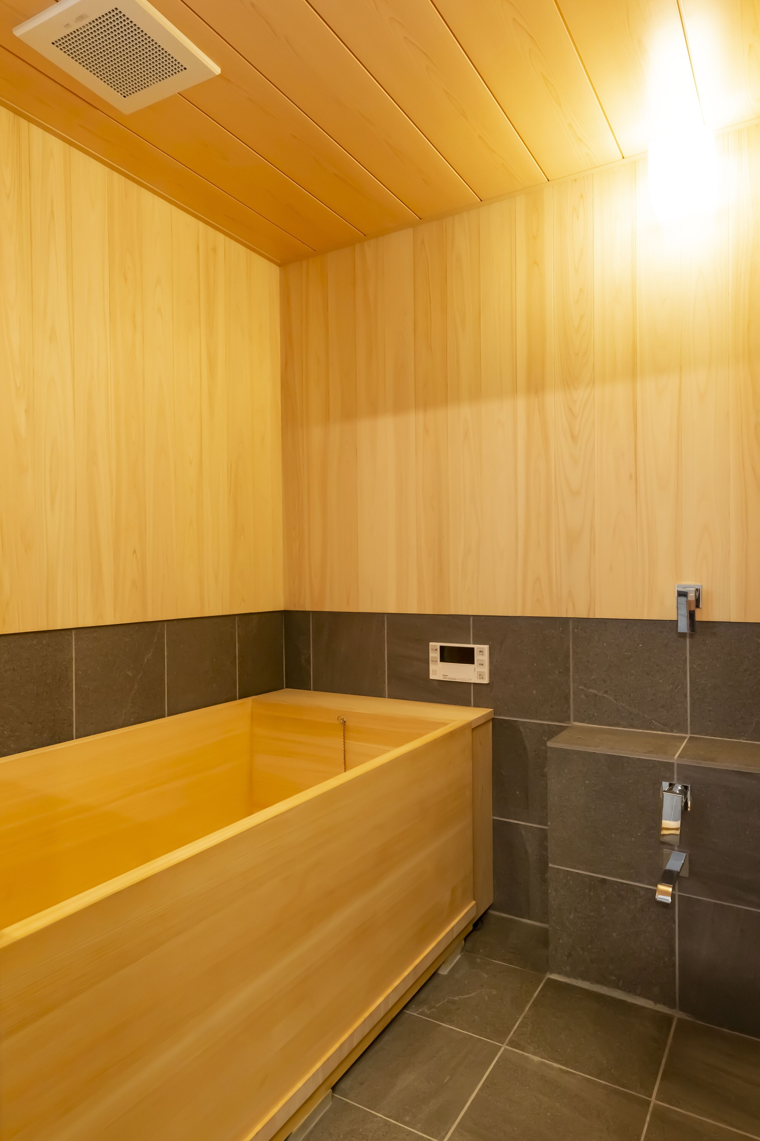 A wooden bathtub in a corner of a room with dark tiled walls and a wooden ceiling.