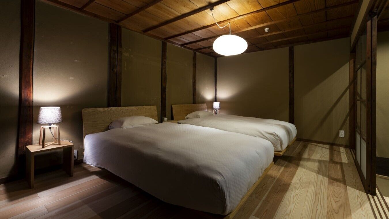 Una habitación con dos camas, piso de madera, una lámpara de techo y una mesita de noche con lámpara.