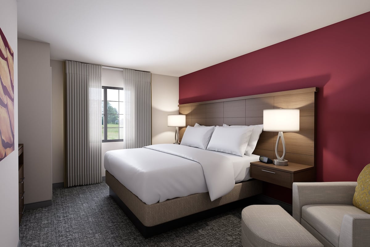 A hotel room with a large bed, two bedside tables with lamps, a chair, and a view of the outdoors through a window with curtains.
