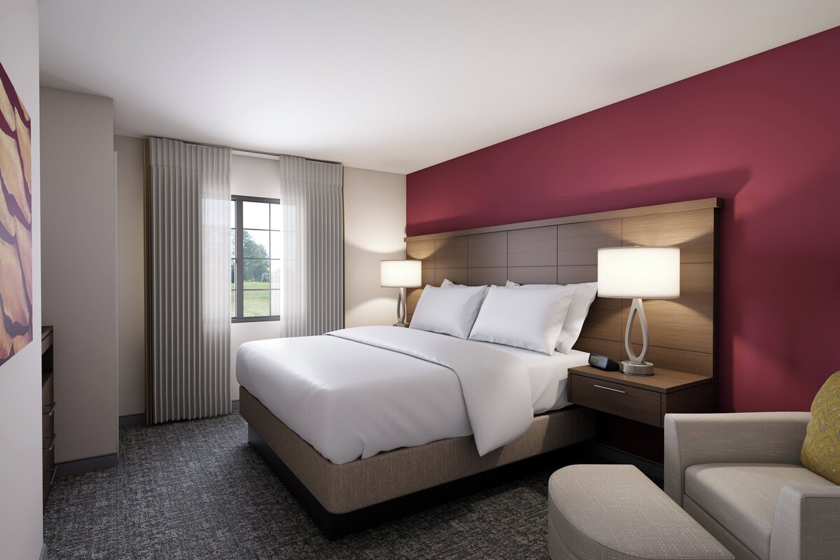 Een hotelkamer met een groot bed, twee nachtkastjes met lampen, een stoel en uitzicht op het omliggende landschap door een raam met gordijnen.
