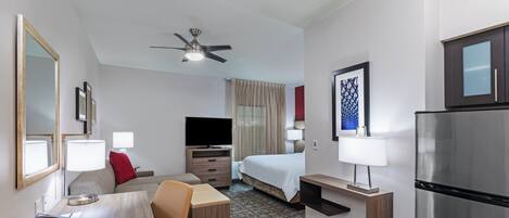 A hotel room with a bed, desk, chair, television, refrigerator, and a ceiling fan.
