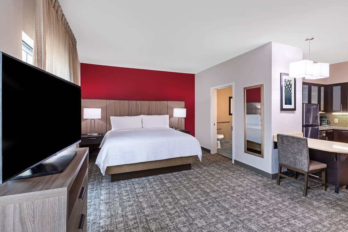 A hotel room with a large bed, a TV, a desk with a chair, and a kitchenette.