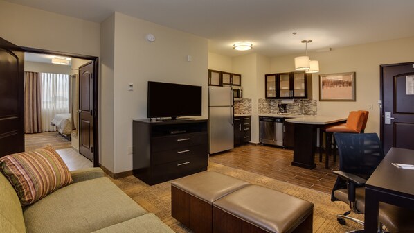 A compact hotel room with a kitchenette, a flat-screen TV, a sofa, an ottoman, and a dining area with a chair.