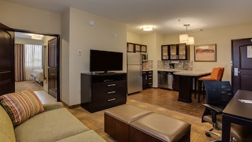 Habitación de hotel compacta con kitchenette, televisor de pantalla plana, sofá, reposapiés y comedor con una silla.