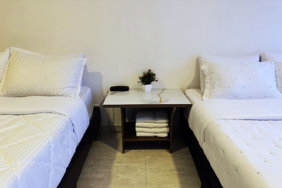 A hotel room with two beds, a small table with a phone and a plant, and a shelf with folded towels.