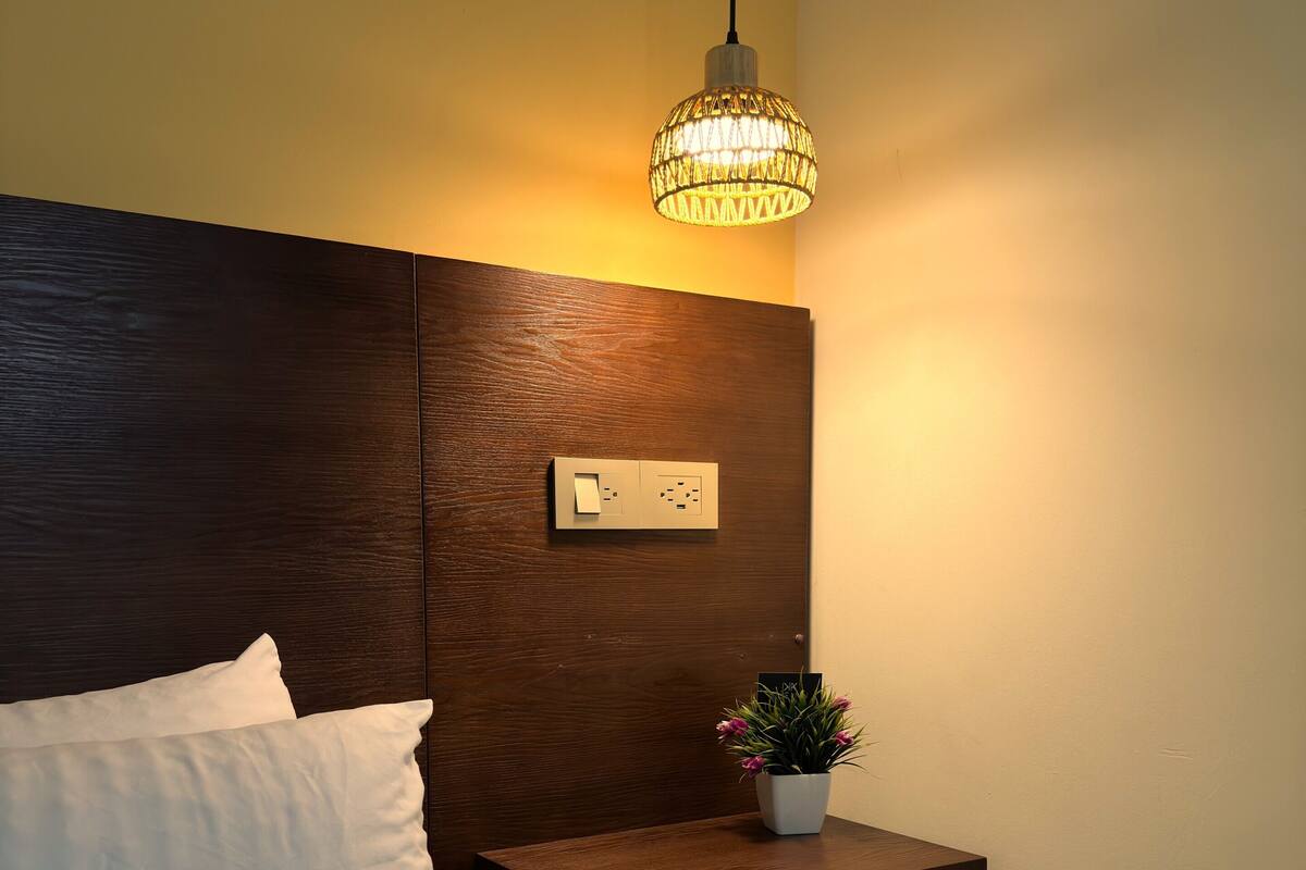 A neatly made bed with white linens, a bedside table with a power outlet, a small potted plant, and a shelf with folded towels.