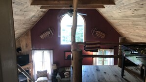 A cozy attic room with a sloped ceiling, wooden beams, a large window, a ceiling fan, a futon, a small table, and a futon bed. - Enjoy private...beautiful… large house with outdoor hot tub and sauna ! (Slaty Fork)