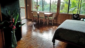 A room with a bed, a dining table with chairs, a vase with flowers, and large windows with a view of greenery. - Evergreen Cottage by Maui Adventure Villa (Haiku-Pauwela)