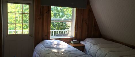 A small, cozy room with two beds, a window with a view of greenery, a ceiling fan, and a door.