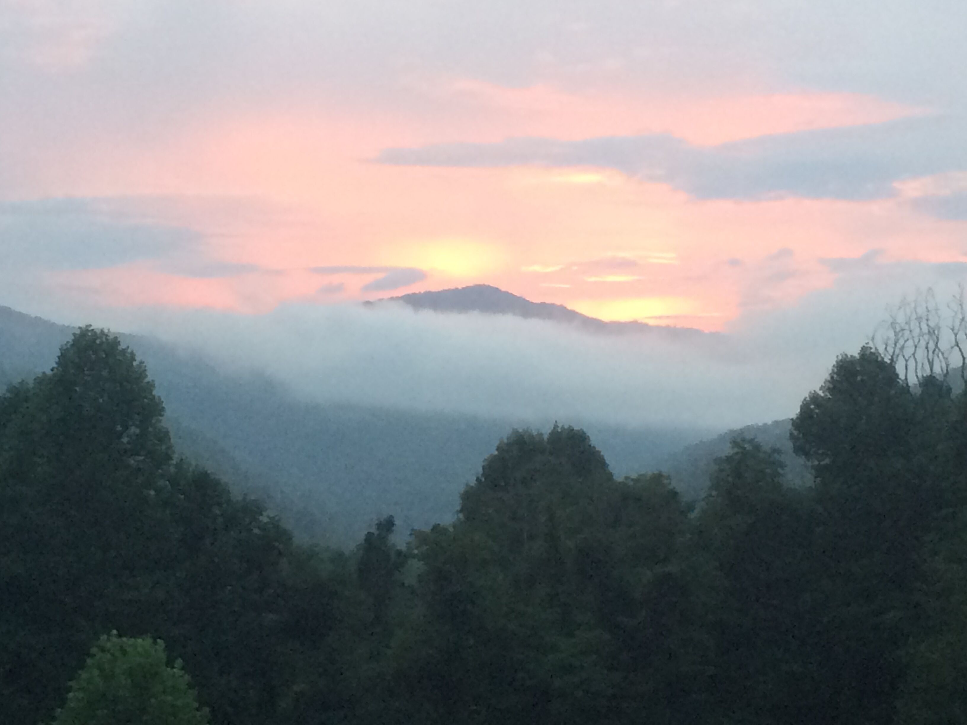 A mountainous landscape with a forested foreground and a sunset in the background.