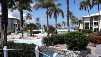 A pool area with lounge chairs, palm trees, and a view of a beach and hotel buildings.