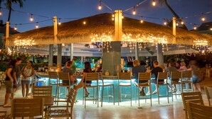Un área de bar al aire libre con piscina, rodeada de sillas y mesas de madera, y luces colgantes.