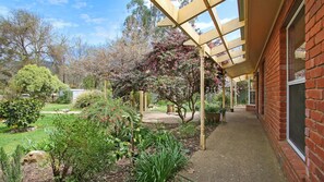 Un espacio al aire libre techado con un edificio de ladrillo, un jardín con diversas plantas y árboles, y un cielo despejado.