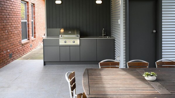 Un área de cocina al aire libre con pared de ladrillo, encimera gris y mesa de madera con sillas.