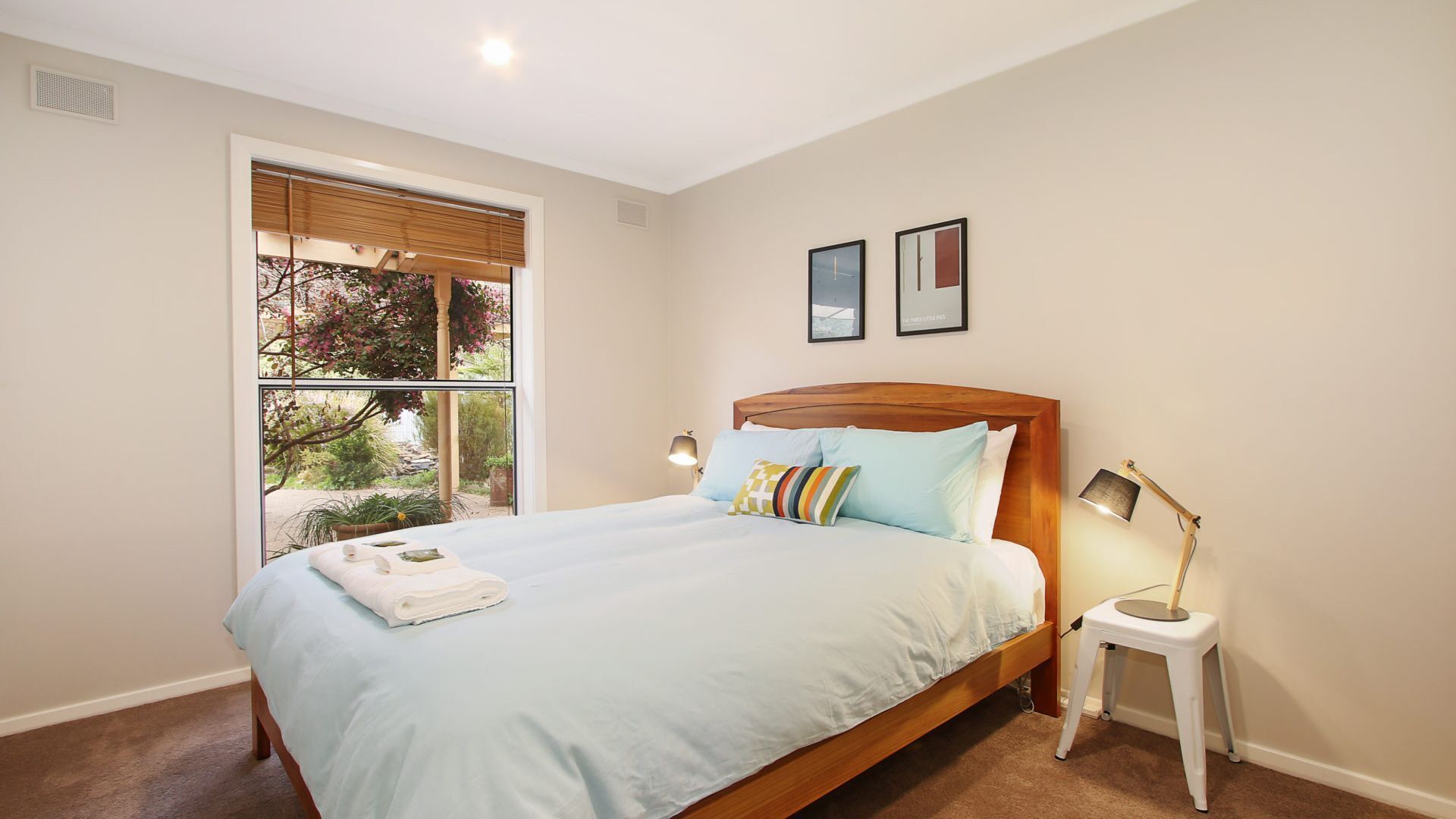 A bedroom with a large bed, a bedside table with a lamp, a window with blinds, and two framed pictures on the wall.