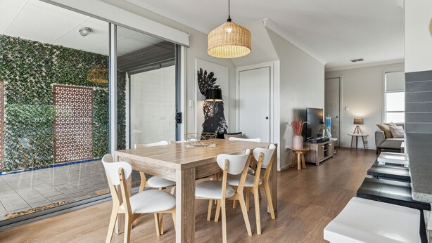 A modern dining area with a wooden table and white chairs, a large woven pendant light, and a view of a lush garden through glass doors.