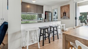 A modern kitchen with white and dark wood cabinets, a stainless steel refrigerator, and a large window overlooking a garden. There are white bar stools and a wooden dining table.