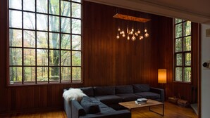 Ein modernes Wohnzimmer mit einem dunkelgrauen Ecksofa, einem Holztisch und einer Pendelleuchte. Große Fenster bieten Blick auf eine bewaldete Gegend.