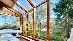 A sunlit room with a large window, wooden beams, a white table, and a cushioned bench.