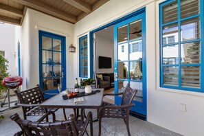 Un área de comedor al aire libre con mesa, sillas, una parrilla y vistas del interior a través de puertas con marco azul.