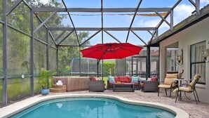 Zona de piscina exterior techada con una sombrilla roja, mobiliario y vistas del entorno verde.