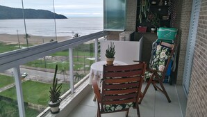 En balkong med ett bord och stolar, en krukväxt och utsikt över stranden och havet.
