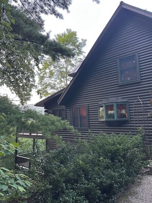 A log cabin with a porch and a small deck, surrounded by dense greenery.