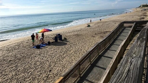 Une plage avec une promenade en bois, des gens sur le sable et un parasol coloré.