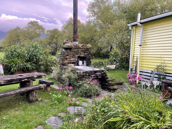 A rustic stone structure with a chimney, surrounded by lush greenery and a yellow building.