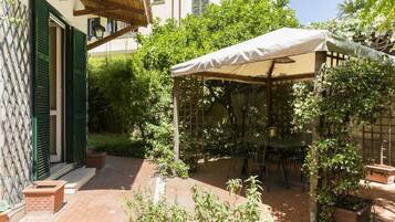Une terrasse avec une table et des chaises, entourée de plantes en pot et agrémentée d’une pergola surmontée d’un auvent blanc.
