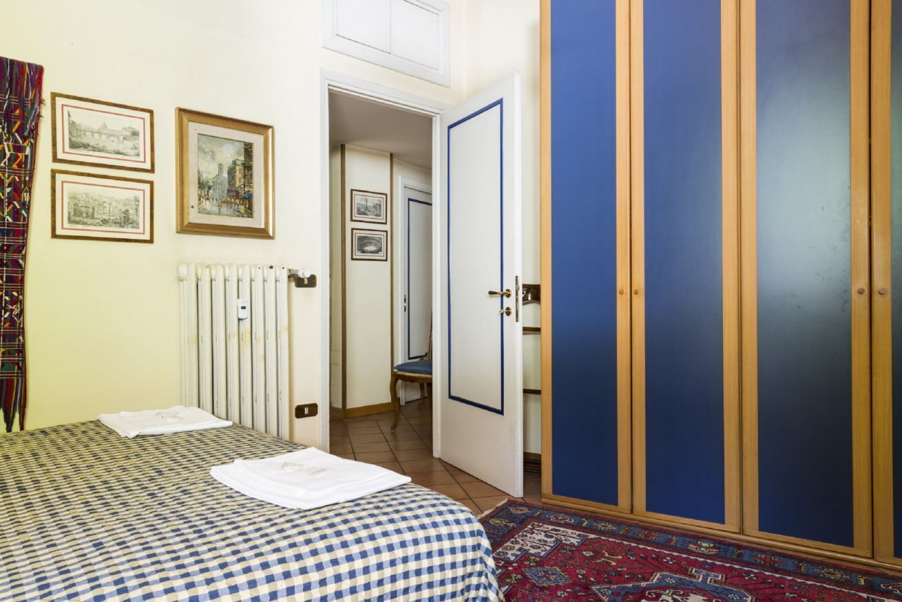 Une chambre avec une couvre-lit à carreaux, un radiateur, des tableaux encadrés accrochés au mur, une armoire bleue et un tapis à motifs.