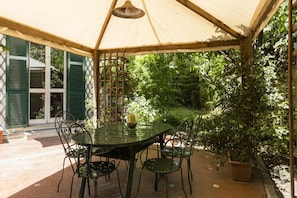Un patio techado con una mesa y sillas, rodeado de follaje.