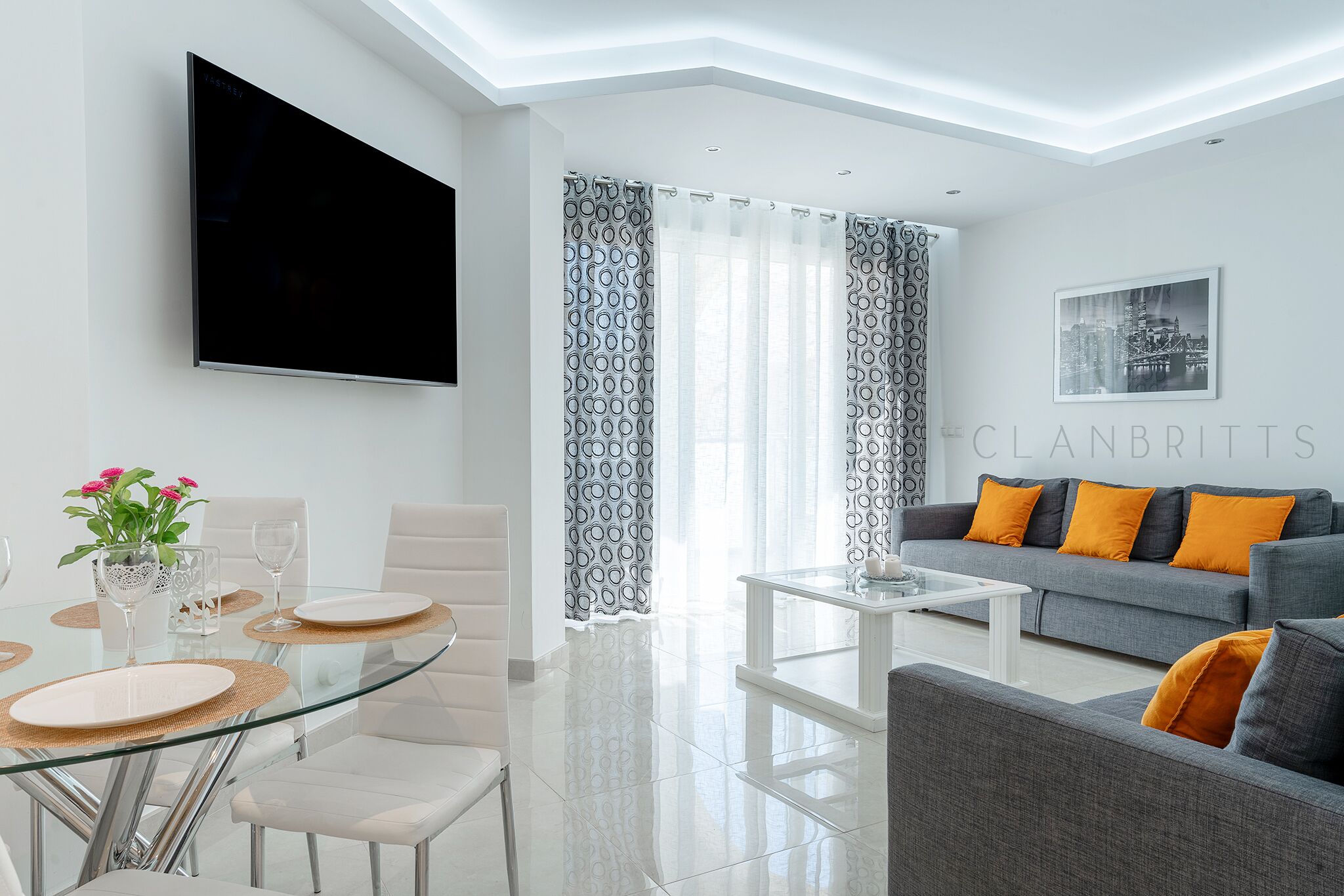 A modern living room with a flat-screen TV, a grey sofa with orange cushions, a glass dining table with white chairs, and a large window with patterned curtains.