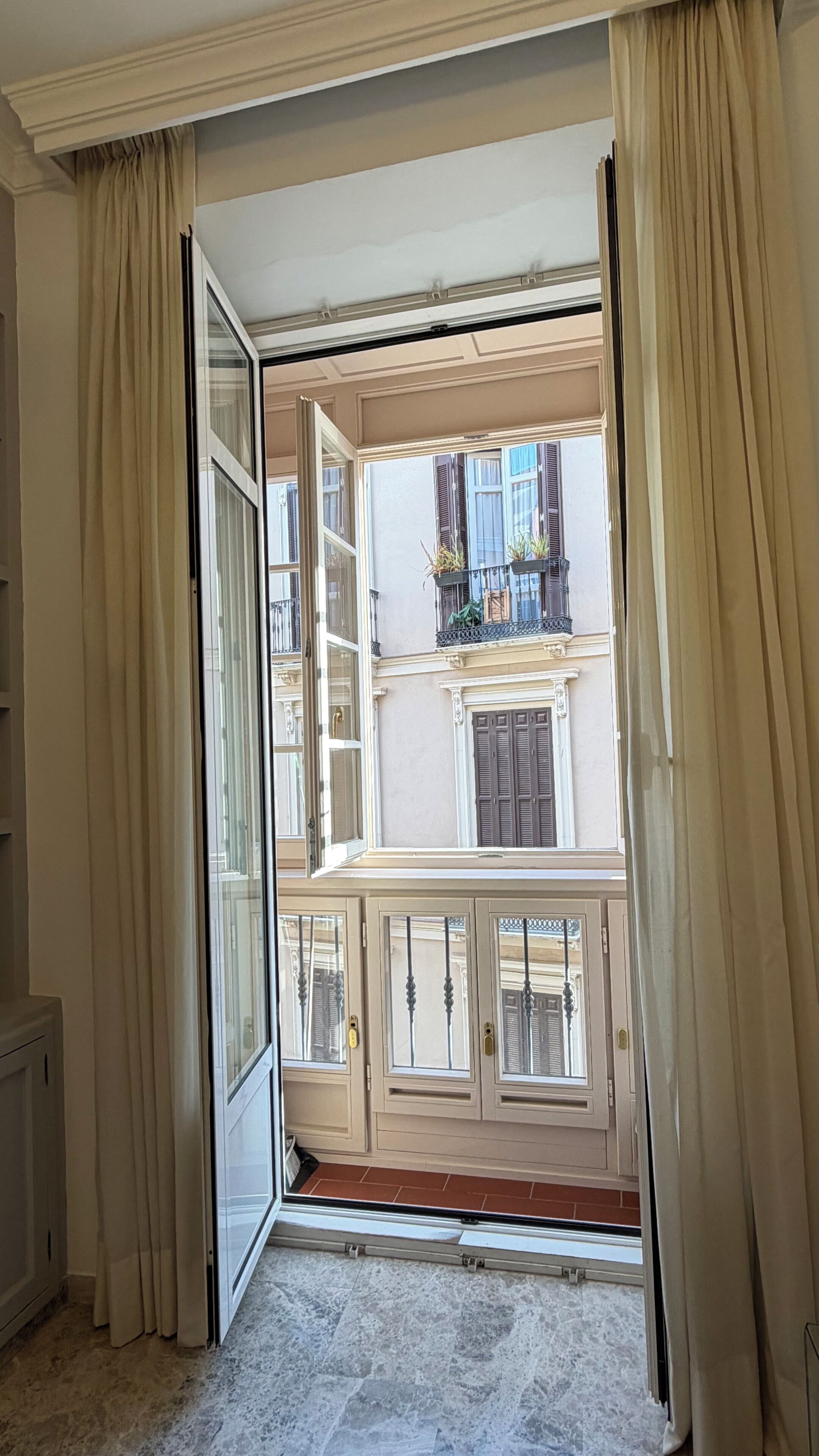 An open sliding glass door leading to a balcony with a view of a building with shutters and a tiled floor.