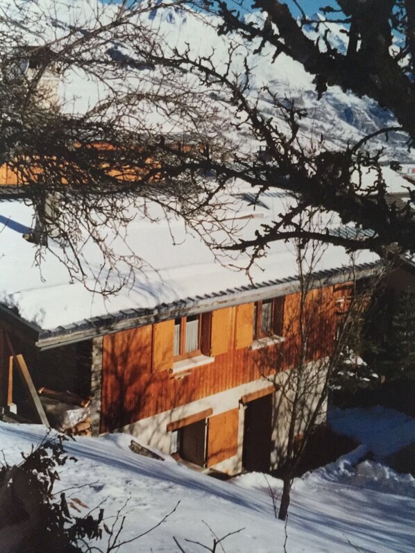 A wooden cabin with a sloped roof, surrounded by snow-covered ground and bare trees.